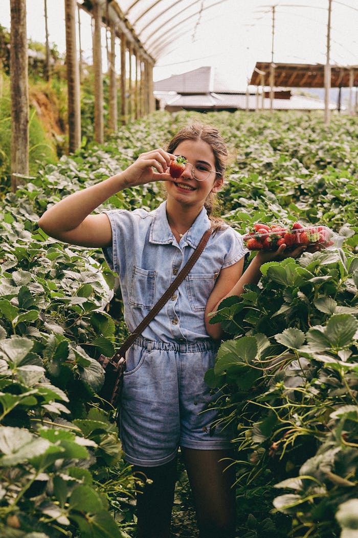 services-04 Young girl in a greenhouse smiling and holding fresh strawberries, surrounded by lush plants.